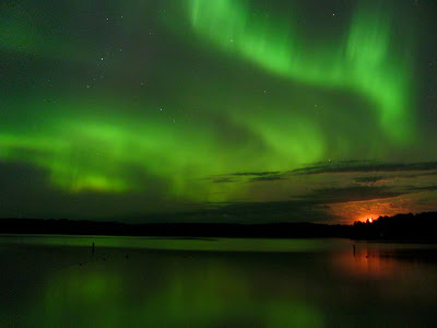 The moon looks orange as it rises. The greenish Aurora Borealis is reflected in a lake.